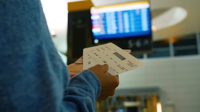 Woman holds passport and boarding pass and looks on departure schedule on screen