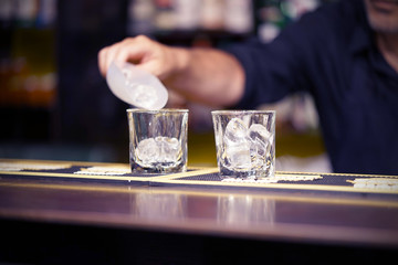 Barman pours ice into a glass. Ice cubes in an empty glass, dark background, front view with details.