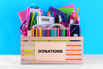 Box with various school and office supplies with a sign on a bright blue background. Donation...