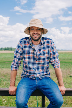handsome middle aged farmer in panama hat sitting on fence and smiling at camera