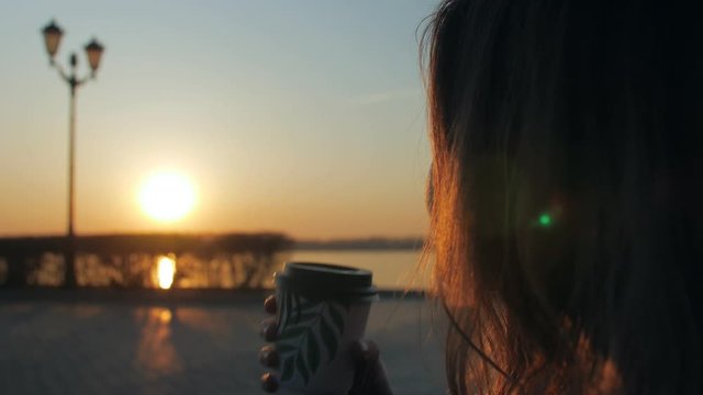 woman sitting on embankment with coffee cup, looking on sun, rising over river in early morning