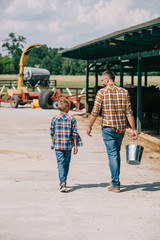 back view of father with bucket and little son walking together on ranch