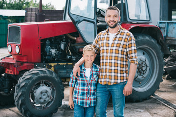 happy father and son in checkered shirts smiling at camera while standing together neat tractor at...