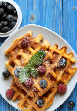 Traditional belgian waffles with fresh berrys and sugar powder on white plate, blue wooden background. Flat lay, top view, copy space.