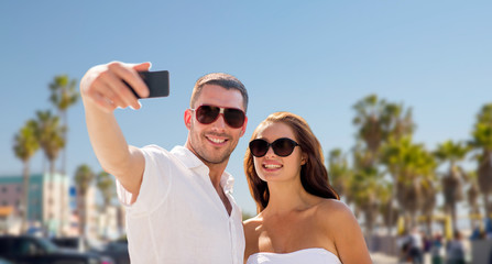 travel, tourism and technology concept - smiling couple in sunglasses making selfie by smartphone over venice beach background in california