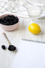 Ingredients for blackberry jam with blank notepad on white wooden surface, side view.