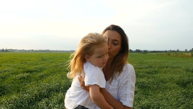 Happy family, mother and her little daughter walk on the green grass, summer evening. Mom with kid walking on green fields. Slow motion