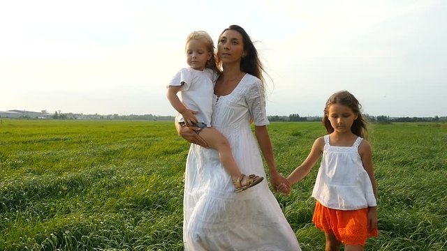 Happy family, mother and her little daughters walk on the green grass, summer evening. Mom with kids walking on green fields. Slow motion