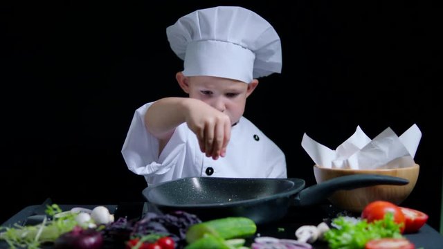 Little boy is adding a salt to the dish, wearing chefs suit and cap. Black background for commercial