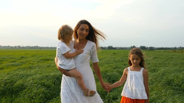 Happy family, mother and her little daughters walk on the green grass, summer evening. Mom with children walking on green fields