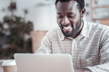 Pleasant job. Happy positive cheerful man looking at the netbook screen and smiling while using it...