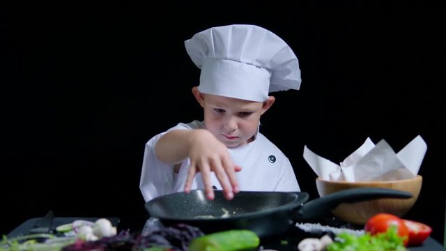 Little boy is adding a salt to the dish, wearing chefs suit and cap. Black background for commercial