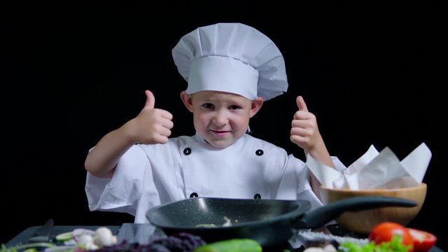 Cheerful boy is dancing and showing thumbs while cooking. He wears chefs suit and cap