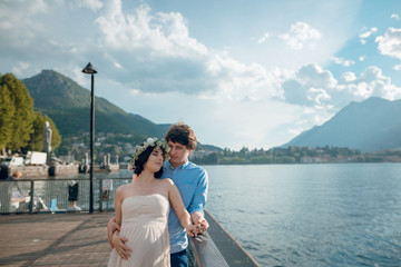 Beautiful young pregnant couple of man and woman standing near the lake Como with scenic mountain view. Sunny summer day. Italy