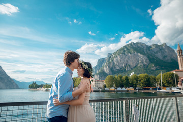 Side view of young family of pregnant woman and her husband standing near the Lake Como on sunny day. Man kissing wife on forehead.