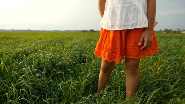 Cute little girl in white-orange clothes standing in green grass. Wind blowing her hair. Summerfield on the sunset. Slow motion