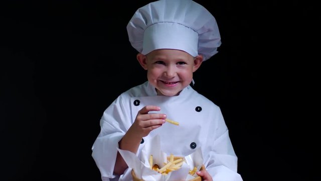 Smily boy wearing chefs suit eats french fries on the black background