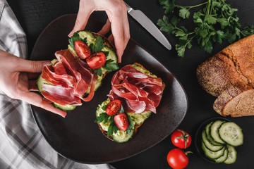 Woman's hands take a sandwich with prosciutto, avocado, cucumber, tomatoes on black stone table, decorated with napkin, knife.