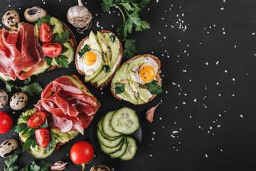 Various of sandwiches and bruschetta with prosciutto, fried quail egg, avocado, cucumber, tomatoes, on black stone background.