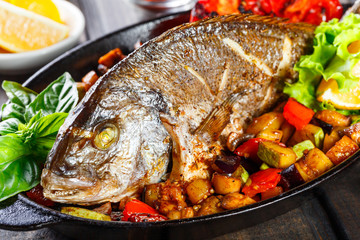 Baked dorado fish with vegetables and lemon on pan on wooden background close up. Delicious dish of seafood