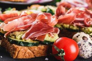 Whole grain bread sandwiches with prosciutto, avocado, cucumber, tomatoes, herbs and seeds on black stone background.