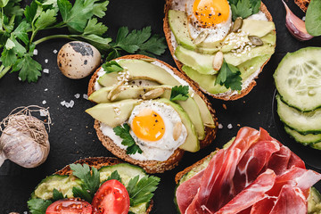 Various of sandwiches and bruschetta with prosciutto, fried quail egg, avocado, cucumber, tomatoes, on black stone background.