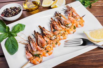 Roasted shrimps on skewers with sauce and lemon on white plate on wooden table. Delicious dish of seafood.