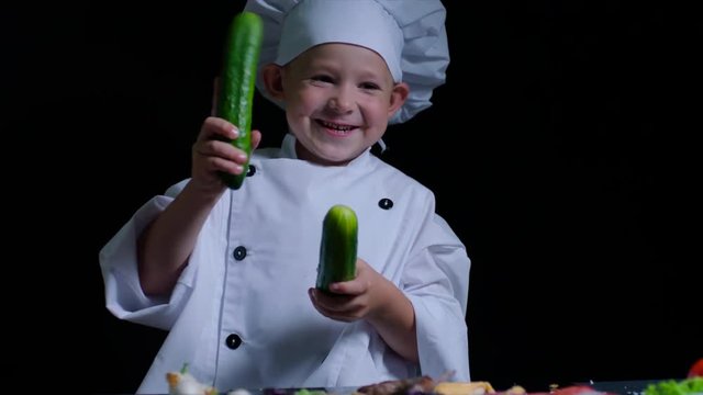 Merry boy wearing chefs suit plays with cucumbers at the cooking table. Black background