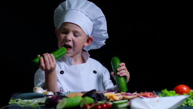 Cute boy bites a cucumber while cooking. He is wearing chefs suit ans cap. Black background