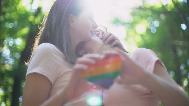 Happy lesbian couple in love kissing, holding rainbow heart, LGBT pride flag