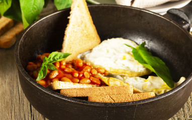 English Breakfast in cooking pan with fried eggs, sausages, bacon, beans, toasts and coffee on dark stone background