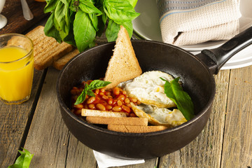 English Breakfast in cooking pan with fried eggs, sausages, bacon, beans, toasts and coffee on dark stone background
