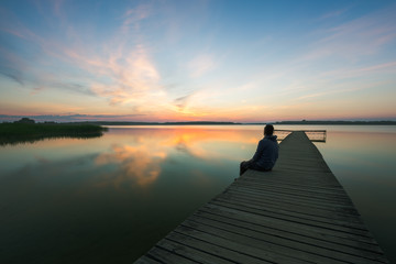 Man is sitting on wooden jetty on lake, during sunrise.