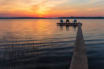 Naklejka na ściany i meble Three empty chairs on wooden jetty on lake, during sunrise.