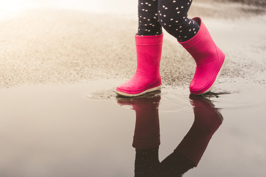 Feet of child in pink rubber boots jumping and splashing over puddle after rain.