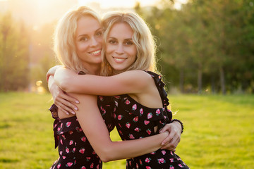 two sisters twins beautiful curly blonde happy young toothy smile women in stylish dress hugging in the summer park sunset rays field background - obrazy, fototapety, plakaty