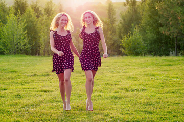 two sisters twins beautiful curly blonde happy young toothy smile women in stylish dress posing in the summer park sunset rays field background