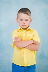 Cute confident serious caucasian little boy in a yellow shirt with arms crossed on a blue background