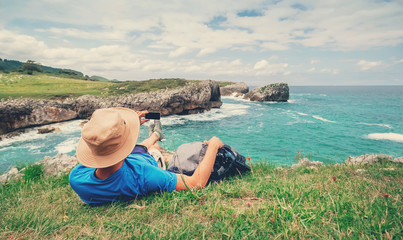 Backpacker traveler rests on the rocky sea side and take mobil photo of beautiful sea landscape