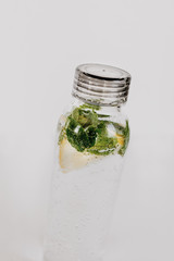 Close up of transparent bottle with water infused with lemon and peppermint leaves to take with on hot summer days. White background, isolated, copy space.