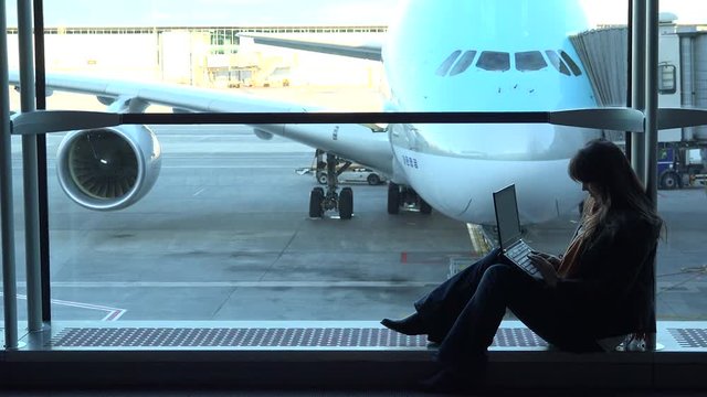 Beautiful woman looking at laptop in airport, sitting at big window, airplane parked