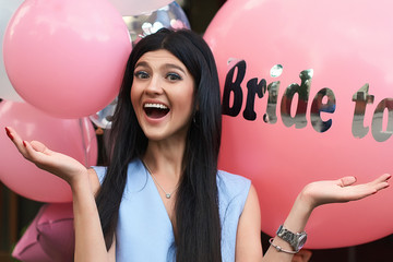 Happy young beautiful brunette bride to be with dark hair and silver crown smiling and gesturing spread out her hands in joy on bachelorette pink party balloon background.