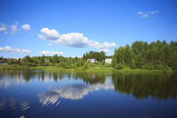Fototapeta na wymiar Lake reflecting sky with clouds on it flowing through a small village and covering banks with trees, shrubs and plants growing on them