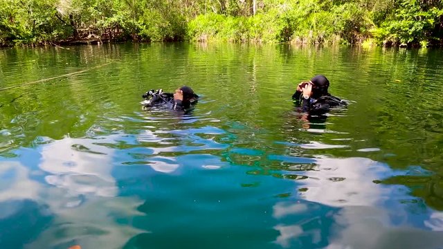 TДва дайвера в полном обмундировании готовятся к погружению в подводную пещеру сенот в Мексике. В зеленой голубой воде, озеро на открытом воздухе против зеленых джунглей.