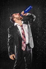 Drunk hopeless young bearded businessman drinking heavily from bottle under the rain, on black background. Social problem concept