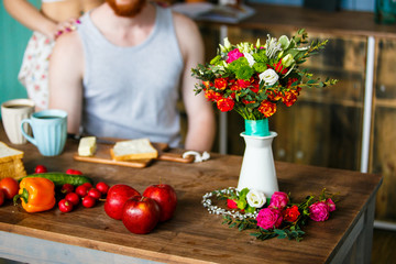 Bouquet of roses in white jug, ripe vegetables on wooden kitchen table. Making breakfast