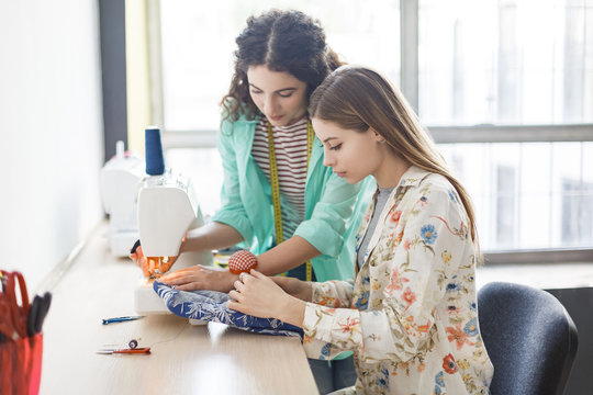 Pretty seamstress teaching girl working with sewing machine at sewing classes with window on background in sewing workshop