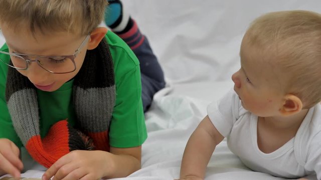 Funny brother lye on belly, bigger brother with eyeglasses read book to attentive baby, happy family portrait, true expression, conceptual