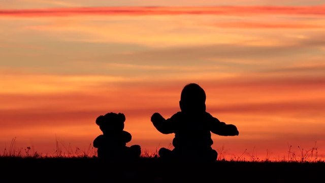 Baby and teddy bear toy silhouette sitting on grass and look at sunset sky