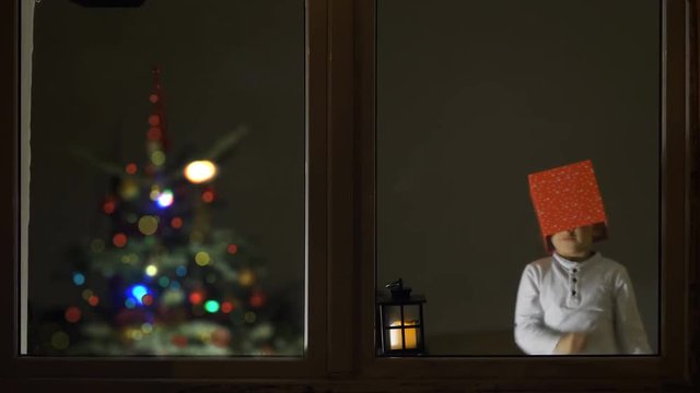 Amusing child fix gift box on his heat, lamp at window, Christmas tree in background, playful and creative childhood